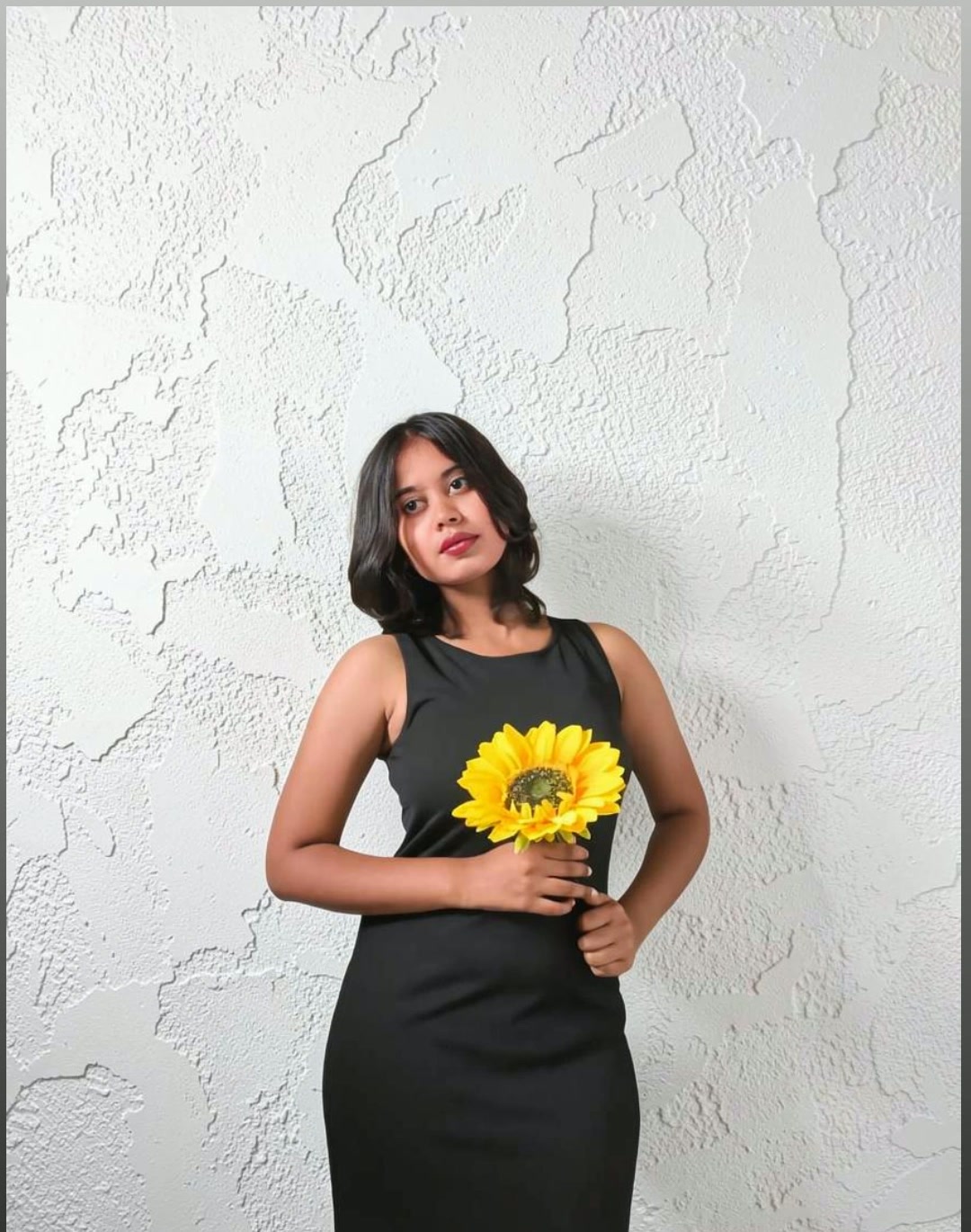 Monisha M in a black dress holding a sunflower on a cream studio backdrop